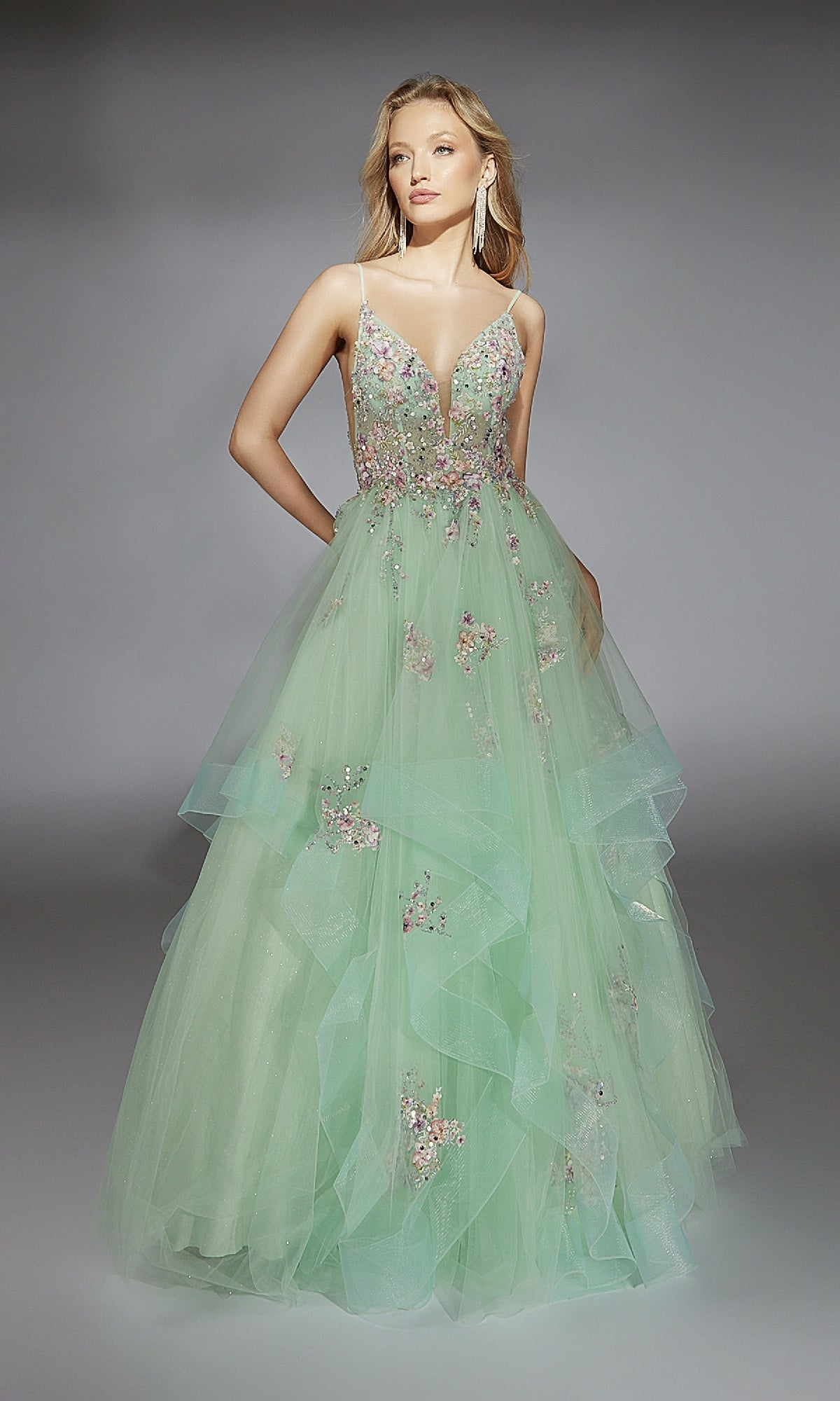 A woman models the Alyce 61735 long prom dress, featuring a light green color, floral embellishments, and a layered tulle skirt, standing against a gray background.